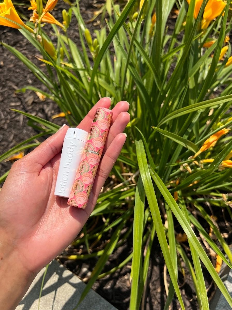 A hand holding lotion buddy beside a lip balm of the same size, in front of yellow flowers